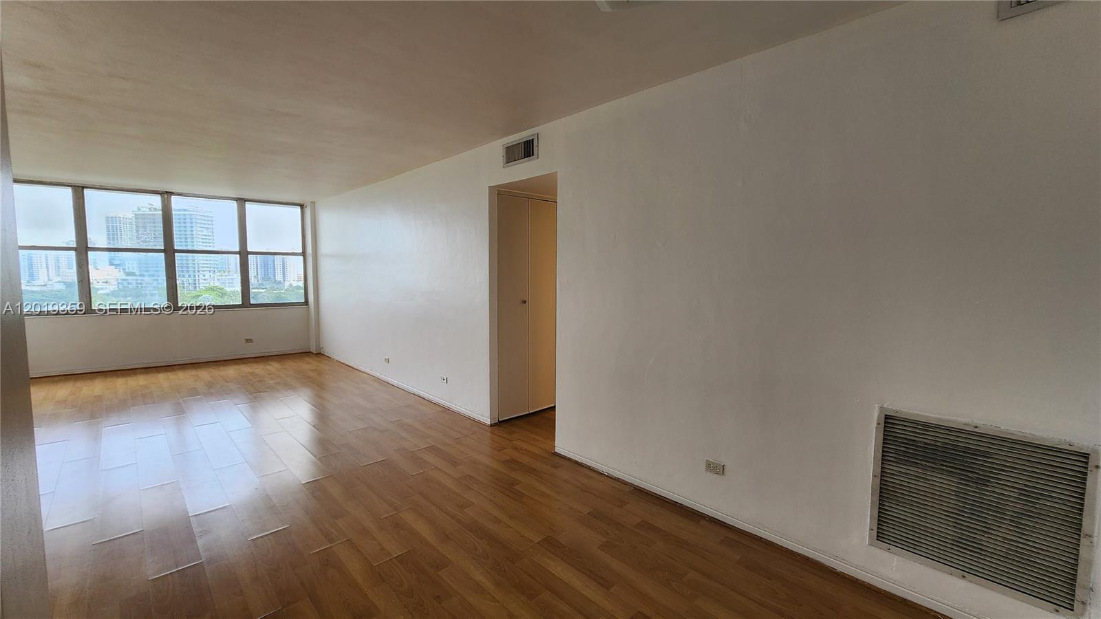 An empty room features light wood flooring, white walls, and a large window with city views — 7441 Wayne # O, Miami Beach FL 33141, MIAMI BEACH, FL