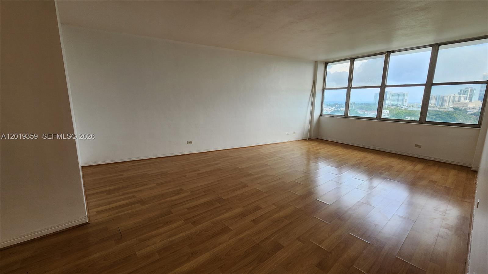 An empty room features light wood-look flooring, white walls, and large windows with city views — 7441 Wayne # O, Miami Beach FL 33141, MIAMI BEACH, FL