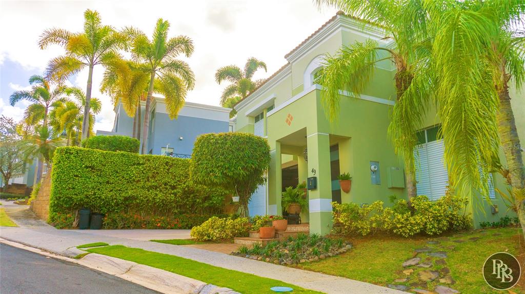 A light green two-story house features a covered entryway, palm trees, and a manicured hedge — 49 Vereda Street MONTEVERDE REAL DEV, SAN JUAN, PR