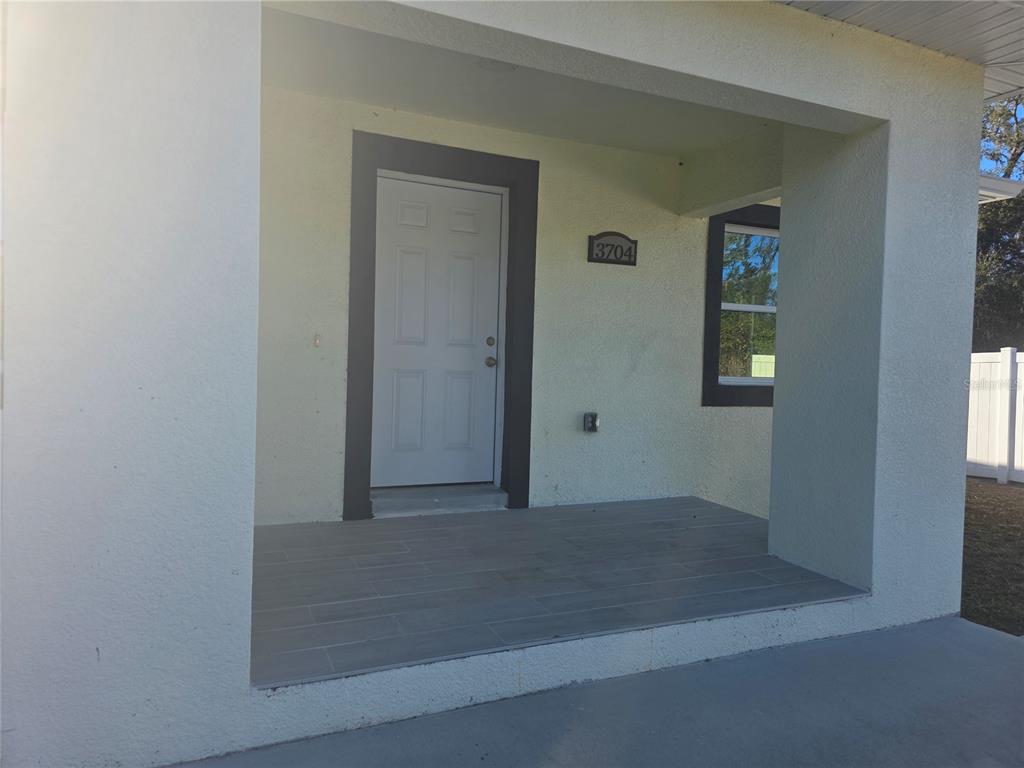 A covered porch features light-colored stucco walls, a white door with dark trim, and tiled flooring — 3704 SMITH TREE STREET, TAMPA, FL