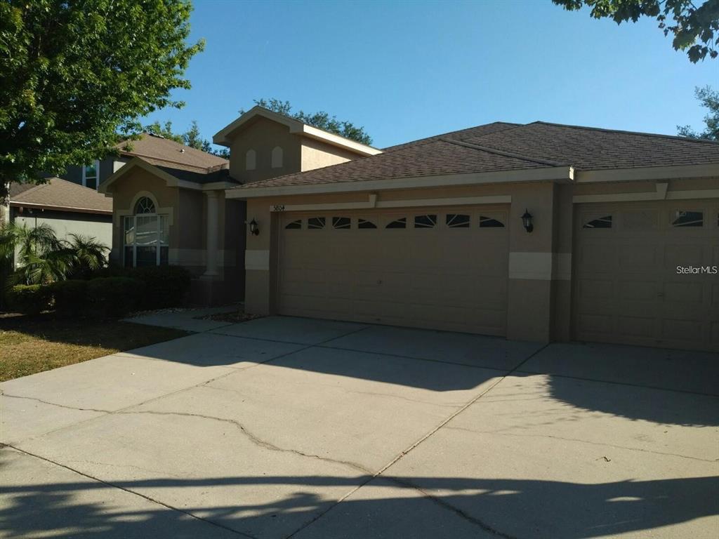 A tan, single-story house features a three-car garage and a concrete driveway — 5804 JUSTICIA LOOP, LAND O LAKES, FL