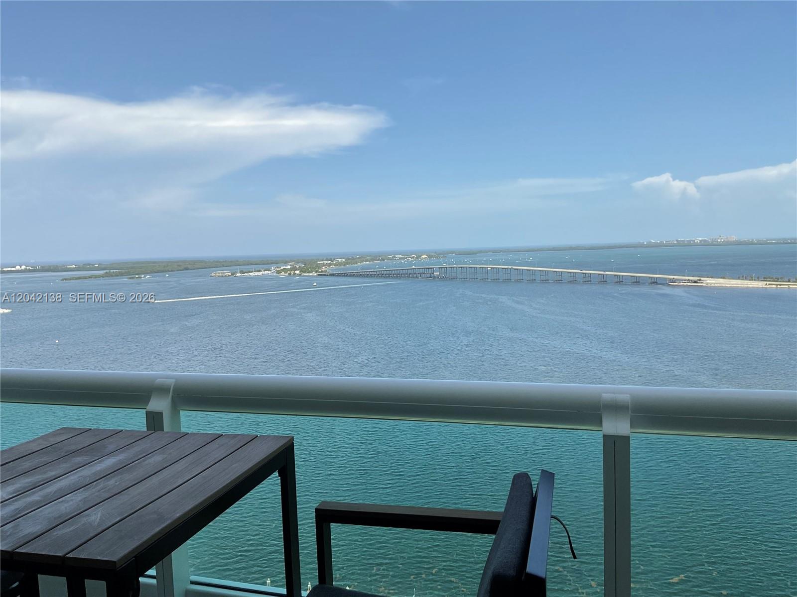 A balcony with a table and chair overlooks a large body of water and bridge — 1643 Brickell Ave # 2603, Miami FL 33129, MIAMI, FL