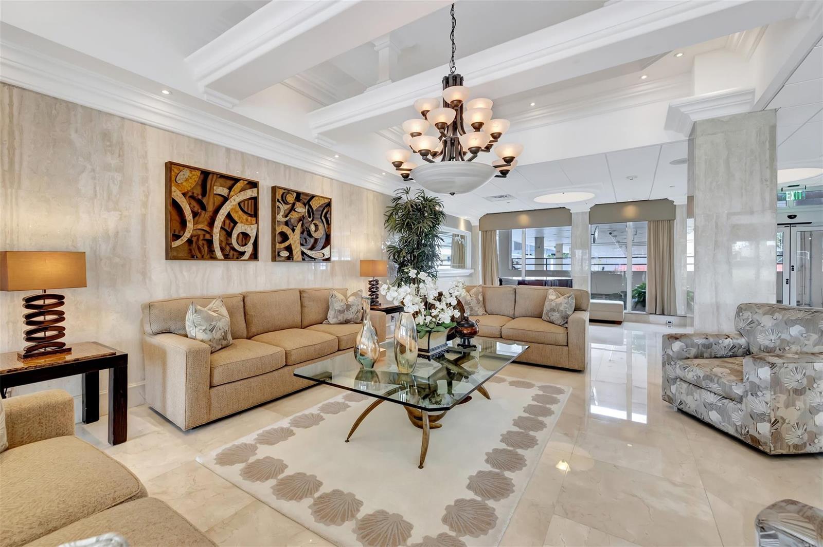 A bright, modern lobby features beige sofas, a glass coffee table, and a large chandelier — 1012 N Ocean Boulevard # 204, Pompano Beach FL 33062, POMPANO BEACH, FL