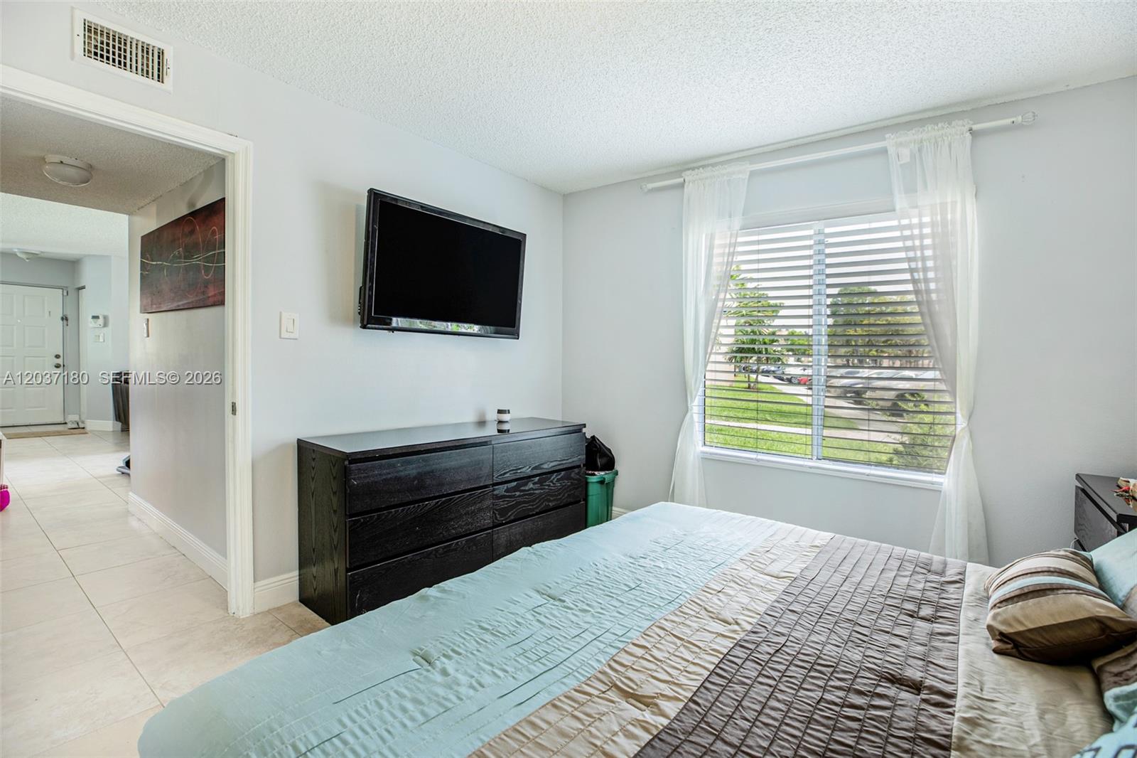 A bedroom features a bed, a black dresser, a wall-mounted television, and a window with blinds — 15780 SW 106th Ter # 107, Miami FL 33196, MIAMI, FL