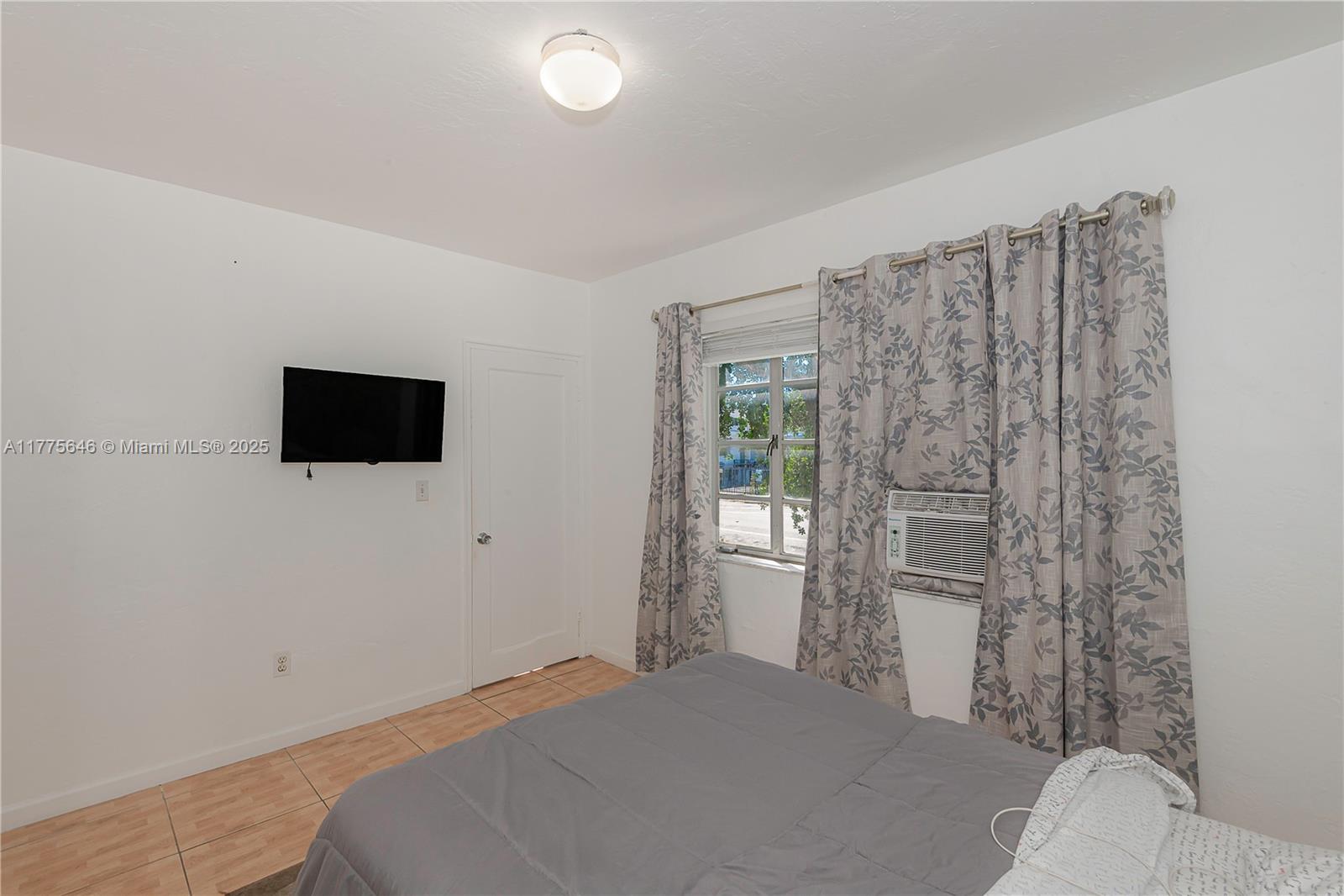 A bedroom features a gray bed, a wall-mounted television, tiled floors, and a window unit — 655 83rd St # 55, Miami Beach FL 33141, MIAMI BEACH, FL