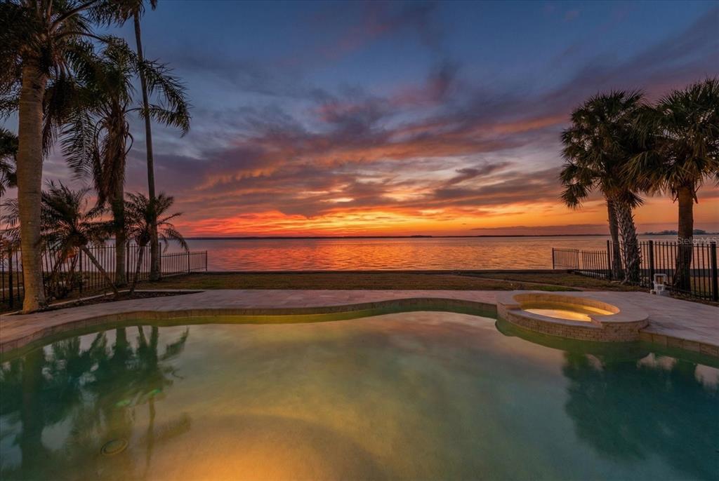 A swimming pool and hot tub overlook a waterfront view at sunset with palm trees — 1614 SANTA BARBARA DRIVE, DUNEDIN, FL