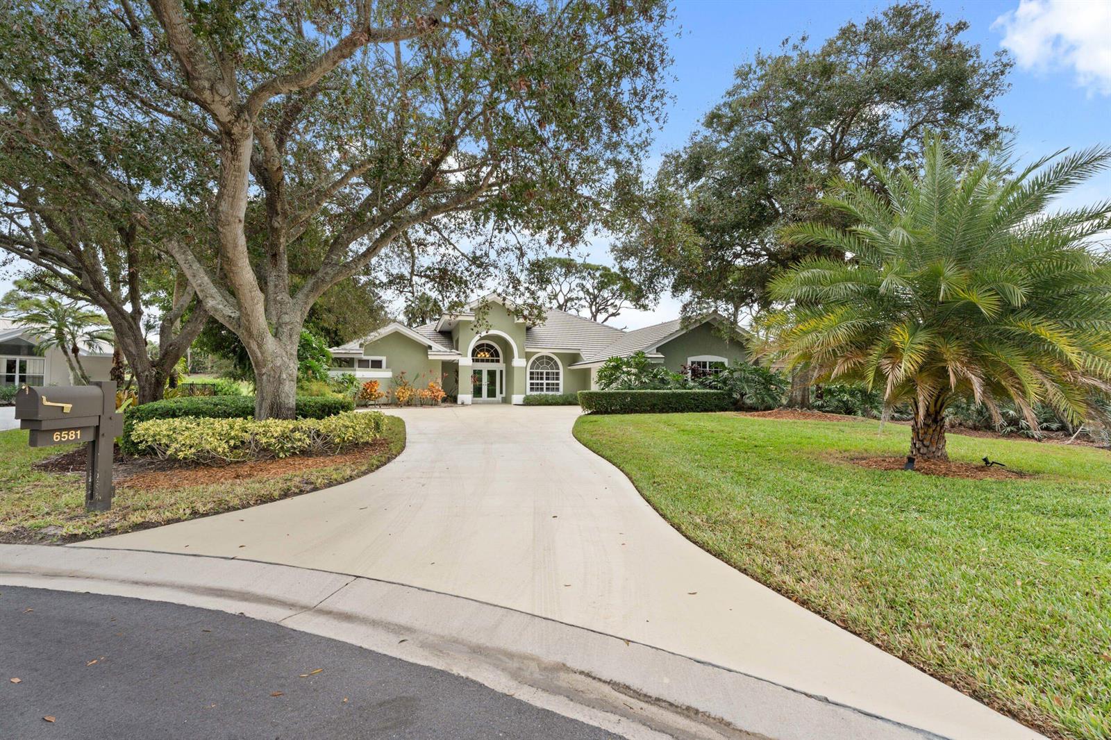 A green house with a curved driveway and large trees sits on a grassy lot — 6581 SE Baltusrol Terrace, Stuart FL 34997, STUART, FL