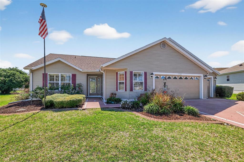 A single-story house with beige siding, a two-car garage, and an American flag — 17559 SE 81ST PARNASSUS COURT, THE VILLAGES, FL