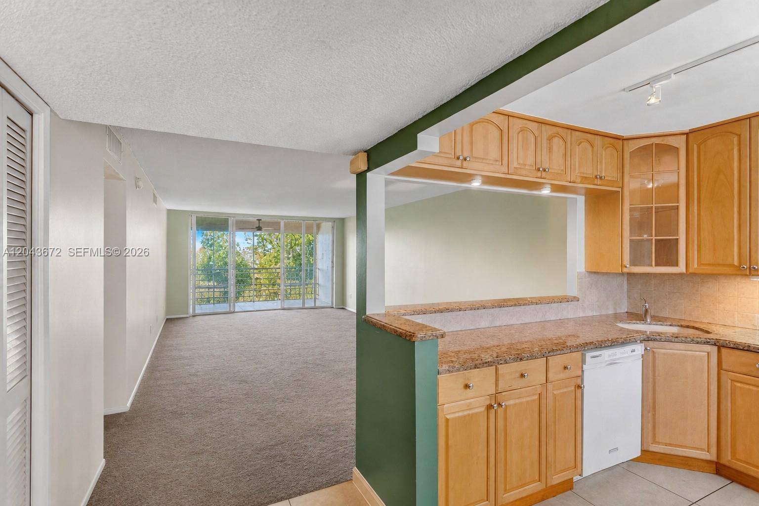 A kitchen with light wood cabinets and granite counters overlooks a carpeted living area — 3010 N Course Dr # 510, Pompano Beach FL 33069, POMPANO BEACH, FL
