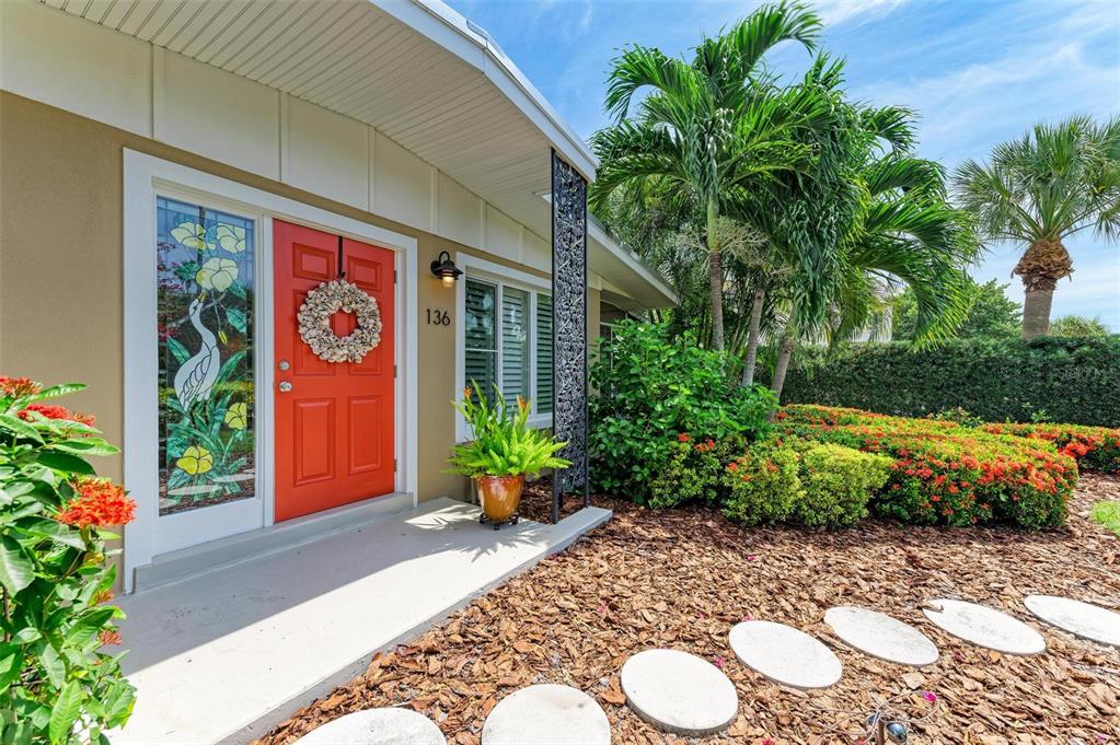 A bright orange front door with a stained glass sidelight sits beside a landscaped walkway — 136 CRESCENT DR, ANNA MARIA, FL