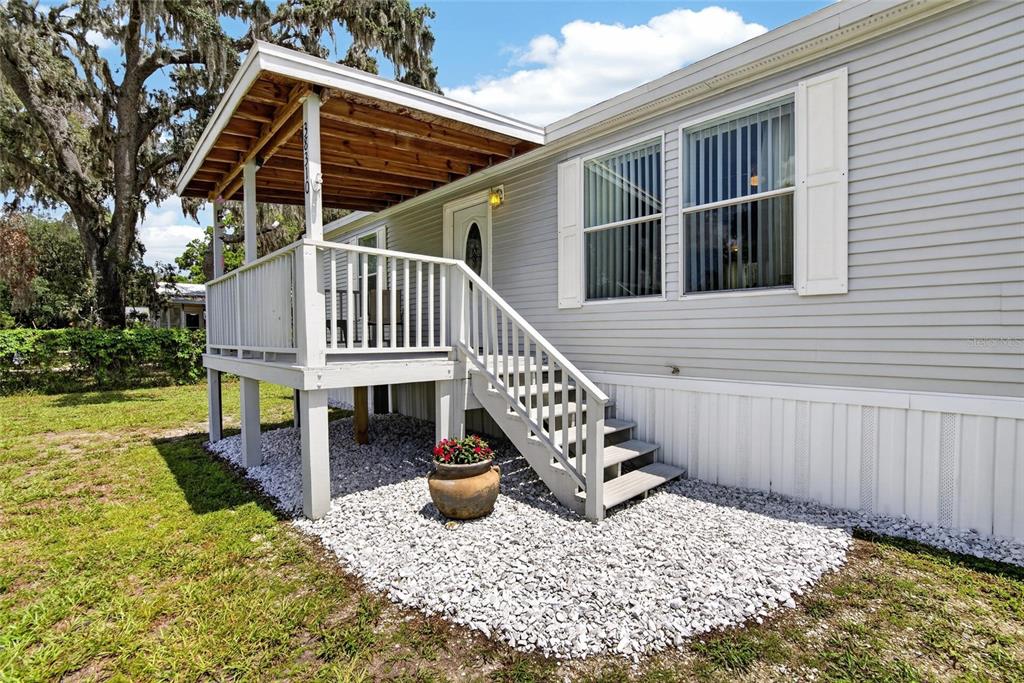 A light gray manufactured home features a wooden front porch with stairs and gravel landscaping — 38310 PEAR COURT, ZEPHYRHILLS, FL