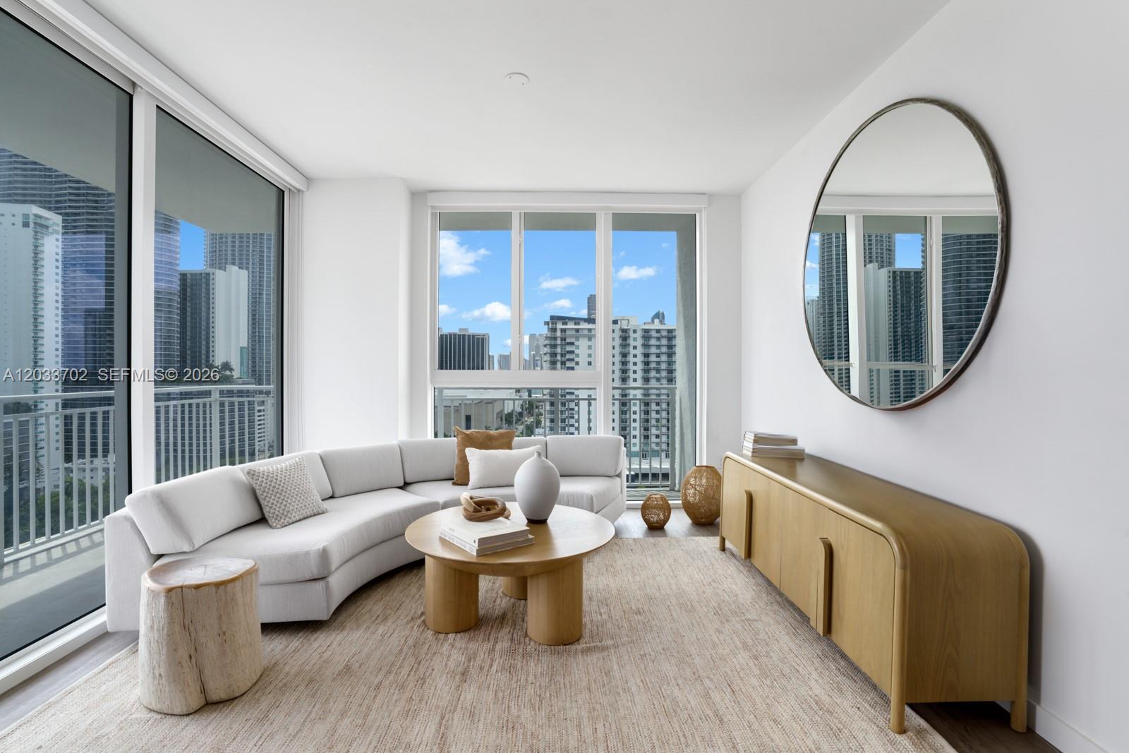A modern living room features a curved white sofa, wooden coffee table, and large round mirror — 2000 Biscayne Blvd # 1707, Miami FL 33137, MIAMI, FL