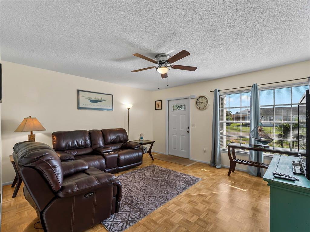 A living room with leather furniture, a ceiling fan, wood parquet flooring, and a front door — 119 OLDENBURGH COURT NE, PORT CHARLOTTE, FL