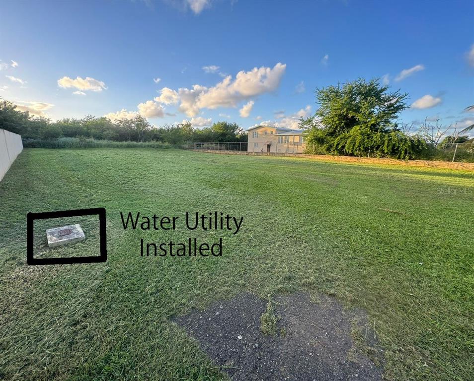 A grassy vacant lot features a marked water utility connection in the foreground — Lot E PR 110 AGUACATE, AGUADILLA, PR