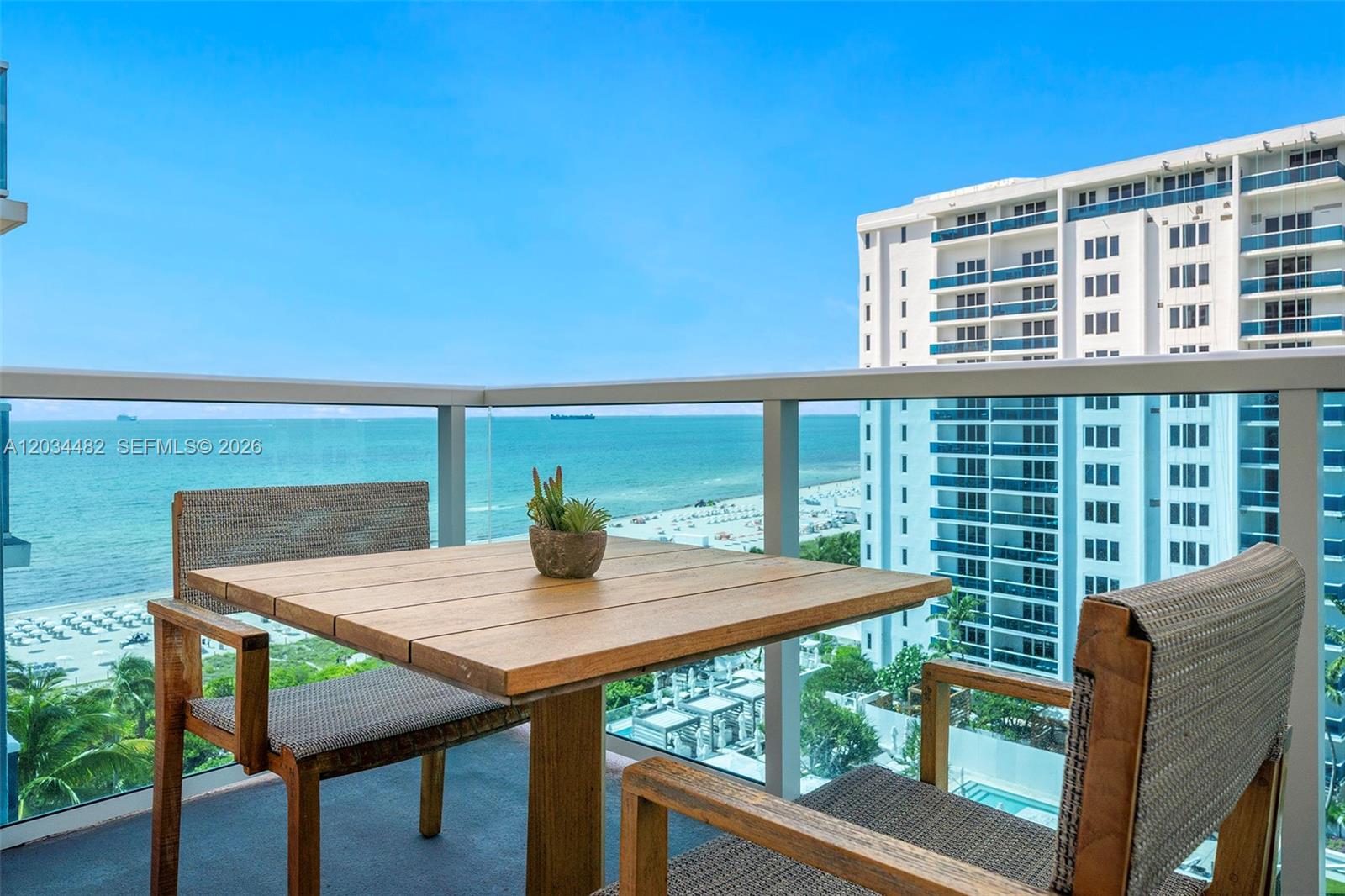 A balcony features a wooden table and chairs overlooking the ocean and a high-rise building — 102 24th St # 1112, Miami Beach FL 33139, MIAMI BEACH, FL