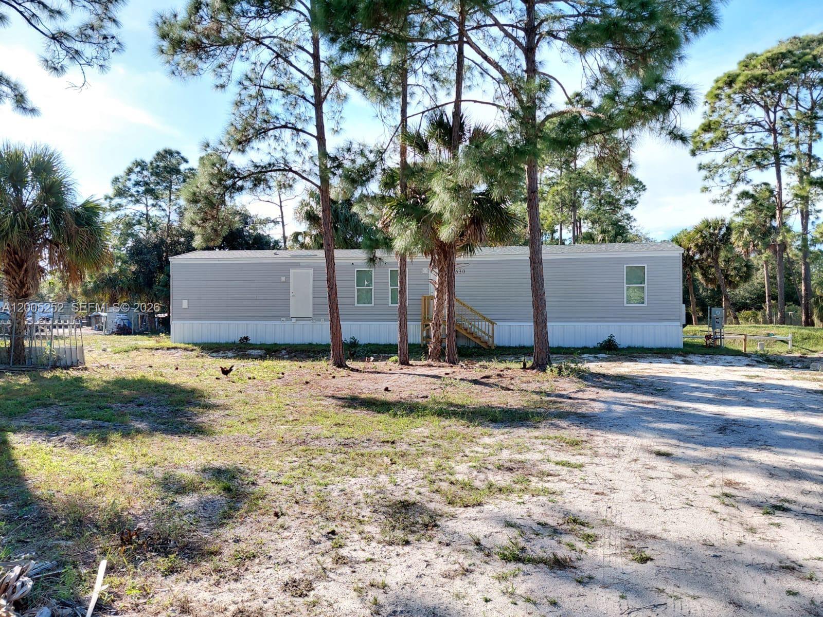 A single-story manufactured home with gray siding sits on a sandy lot among pine trees — 630 S Trebol Street, Clewiston FL 33440, CLEWISTON, FL