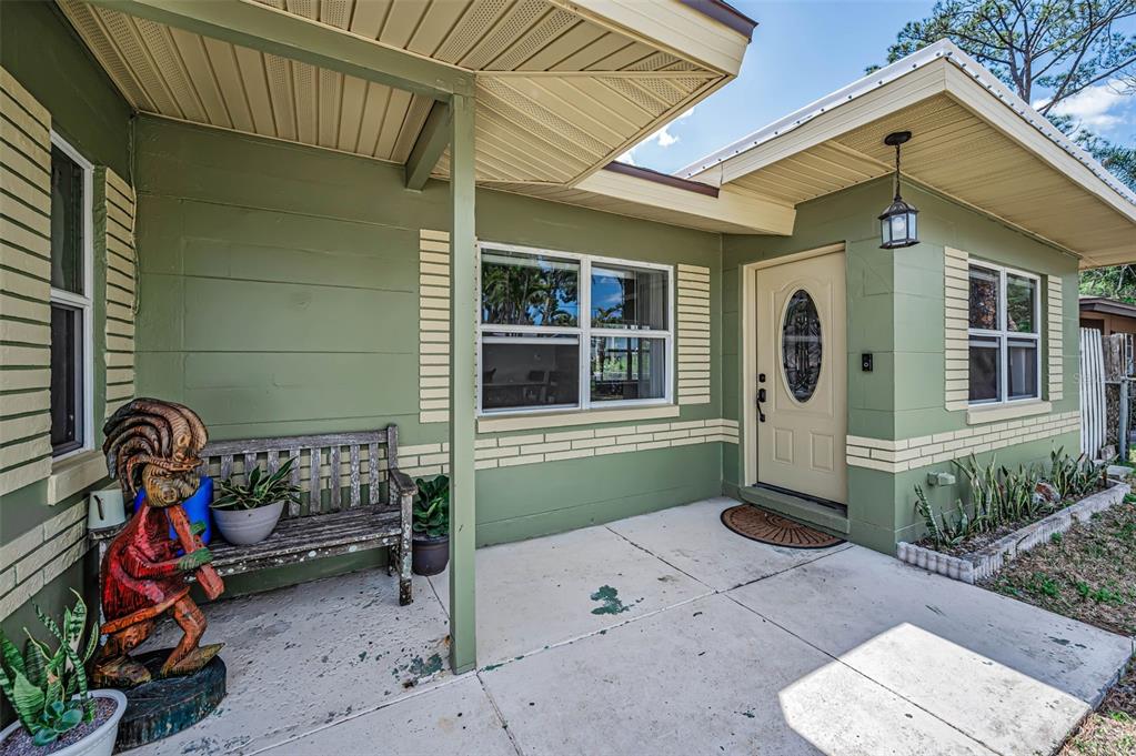 A green house exterior features a covered concrete porch, a wooden bench, and a tiki statue — 5146 MALLETT DRIVE, PORT RICHEY, FL