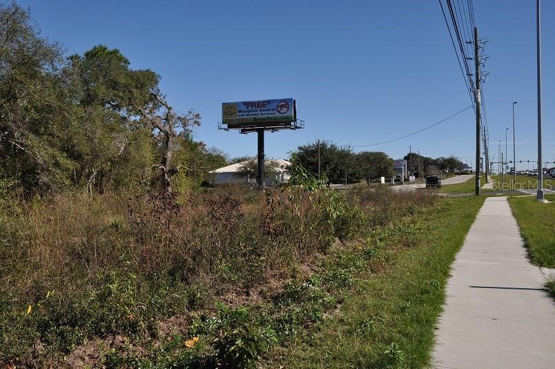 A sidewalk runs alongside a grassy, undeveloped lot featuring a large billboard under a clear sky — 11507 CORTEZ BOULEVARD UNIT 102, BROOKSVILLE, FL