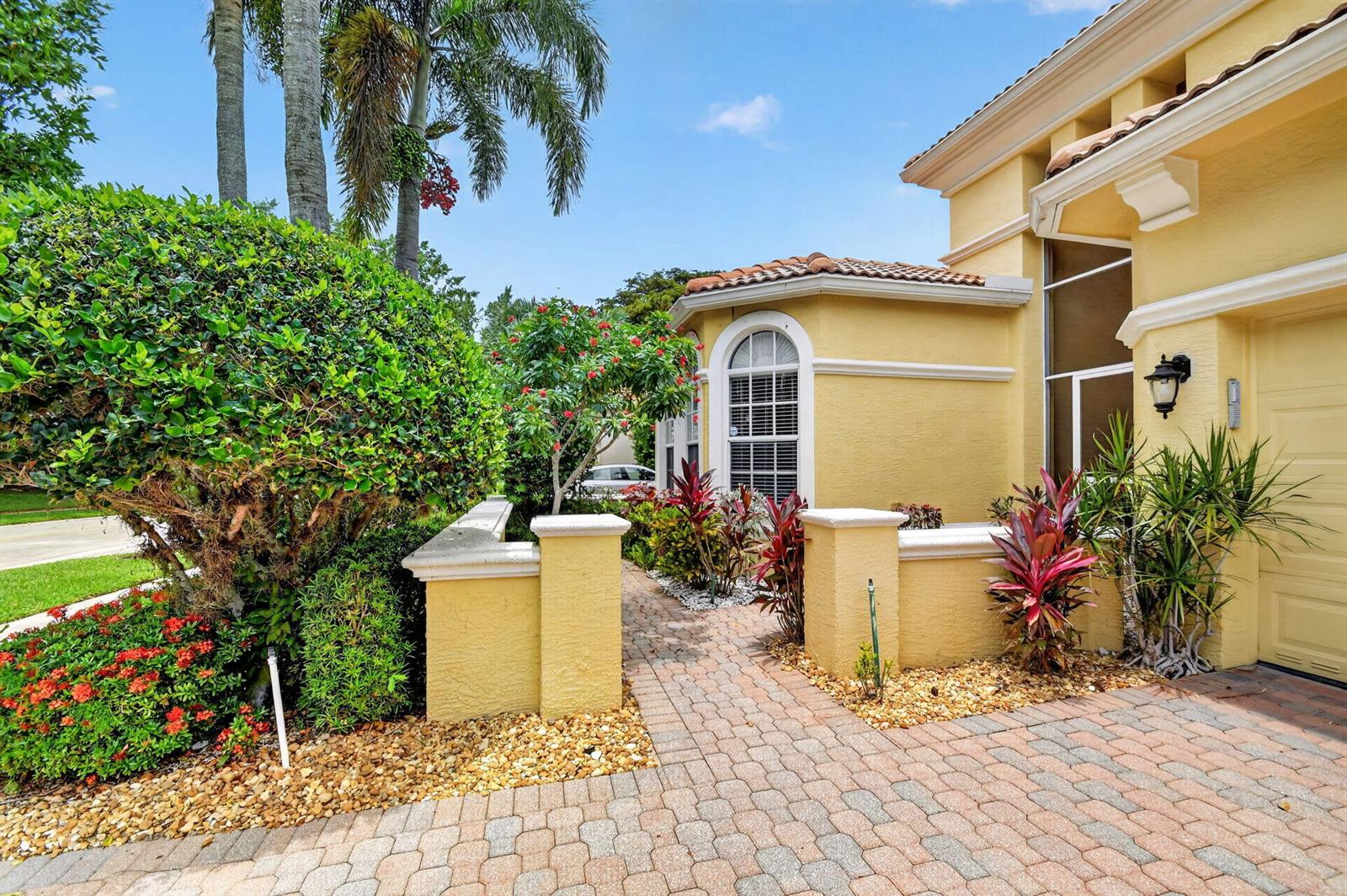 A yellow stucco house exterior features a brick walkway, tropical landscaping, and a garage door — 1848 Via Castello, Wellington FL 33411, WELLINGTON, FL