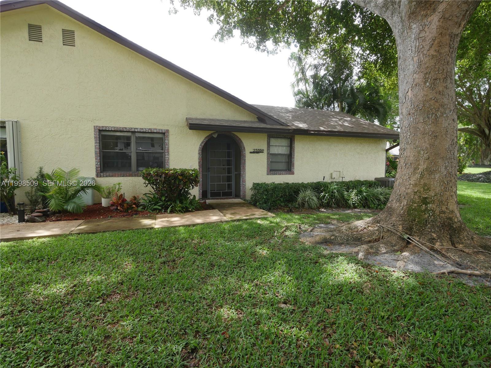A yellow stucco house features a brick-trimmed arched entryway and a large shade tree — 23359 Barlake Dr, Boca Raton FL 33433, BOCA RATON, FL