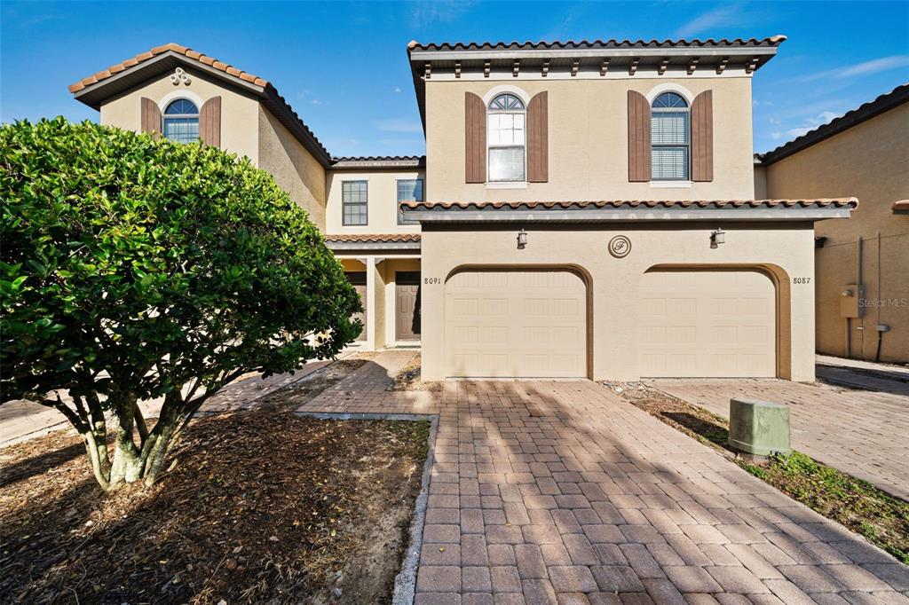A two-story tan stucco townhouse features a paver driveway and two attached garage doors — 8091 BROOKSVILLE DRIVE, DAVENPORT, FL