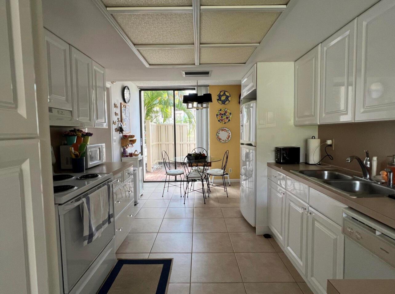A galley kitchen with white cabinets and tile floors leads to a dining area with patio access — 1002 Bridgewood Place # 1002, Boca Raton FL 33434, BOCA RATON, FL
