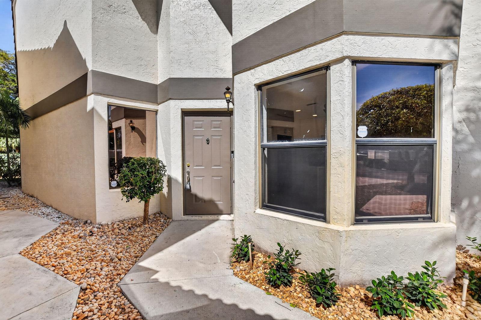 The exterior of a home features a tan front door, bay windows, and landscaping — 6647 Boca Hermosa Lane, Boca Raton FL 33433, BOCA RATON, FL