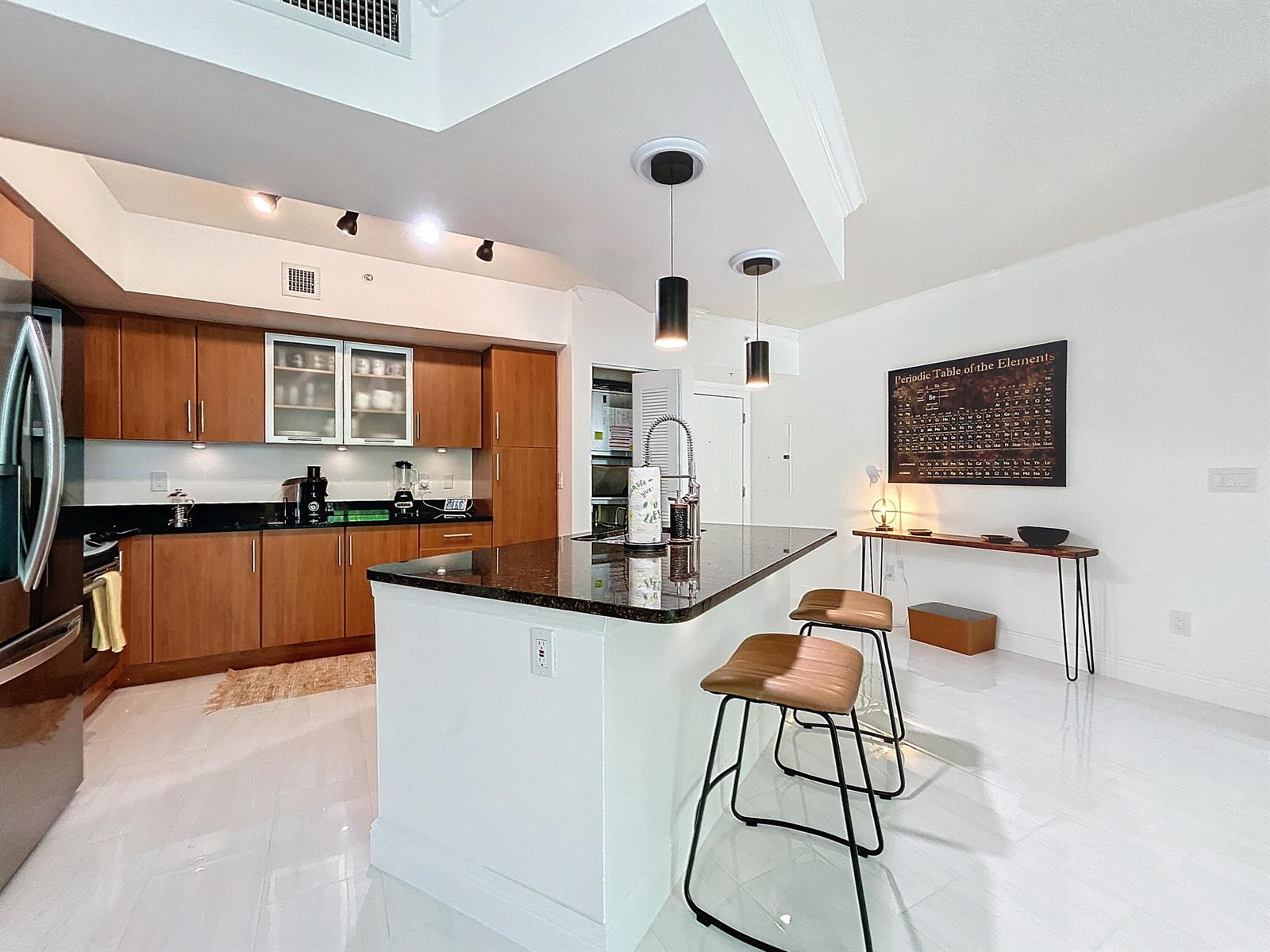 A modern kitchen features wood cabinets, a dark granite island with bar stools, and white flooring — 600 S Dixie Highway # 630, West Palm Beach FL 33401, WEST PALM BEACH, FL