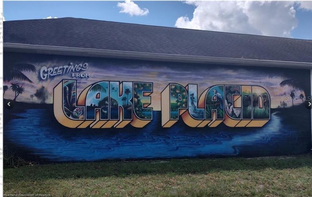 A large mural on the exterior wall of a building reads Greetings From Lake Placid — 123 PATTON AVENUE, LAKE PLACID, FL