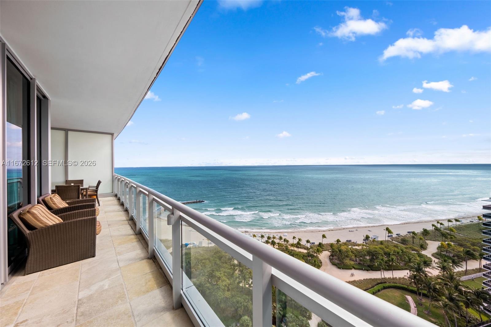 A balcony with wicker furniture overlooks the ocean, sandy beach, and palm trees — 10295 Collins Ave # 1605, Bal Harbour FL 33154, BAL HARBOUR, FL