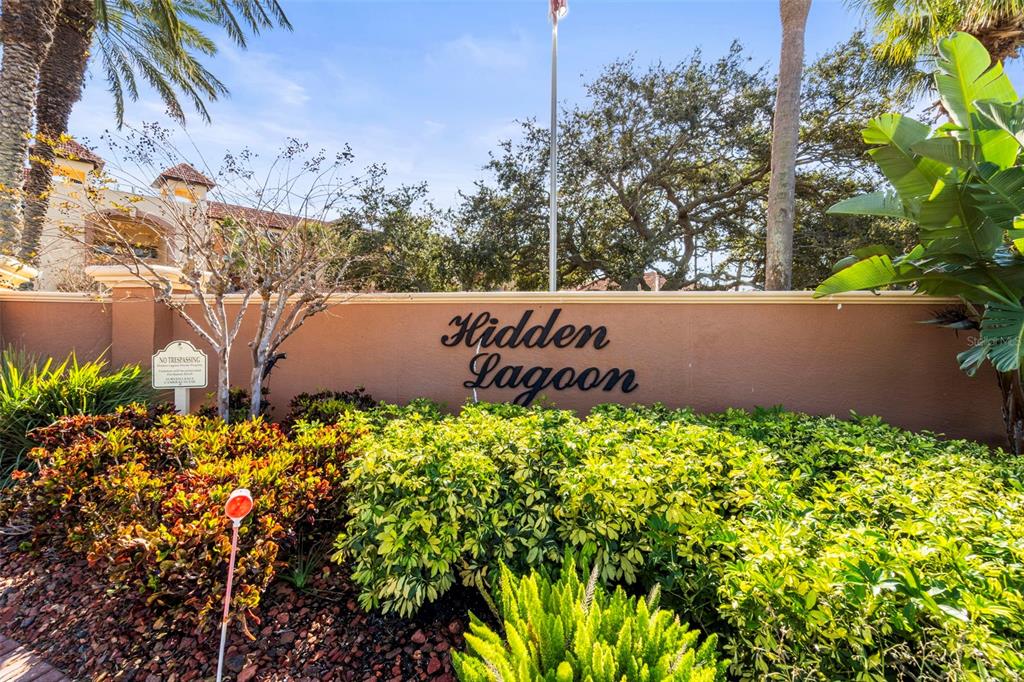 A tan wall with the words Hidden Lagoon stands behind a garden of green shrubs — 1645 PINELLAS BAYWAY S UNIT B2, TIERRA VERDE, FL
