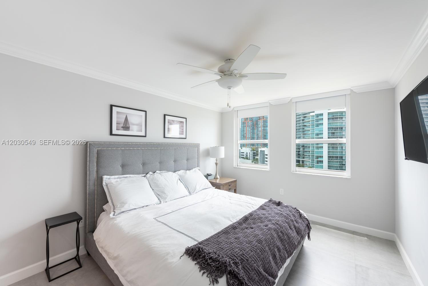 A bedroom features a gray tufted headboard, a ceiling fan, and two windows overlooking buildings — 90 Alton Rd # 1102, Miami Beach FL 33139, MIAMI BEACH, FL