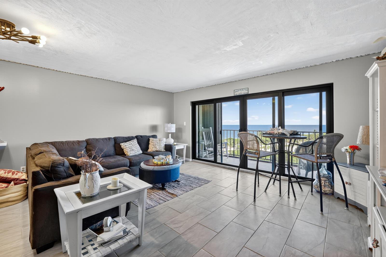 A living room features a dark sectional sofa, tile flooring, and sliding glass doors overlooking the ocean — 801 S Ocean Drive # 910, Fort Pierce FL 34949, FORT PIERCE, FL