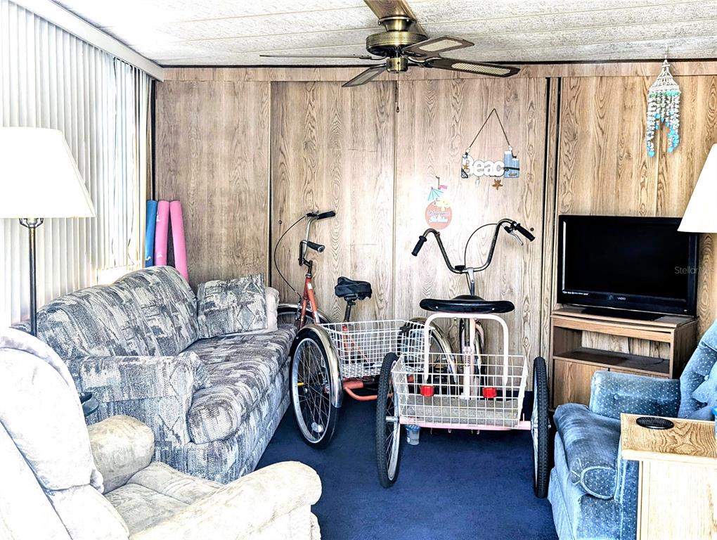 A living room features wood-paneled walls, blue carpeting, two adult tricycles, and patterned furniture — 3300 26TH AVENUE E UNIT 228, BRADENTON, FL