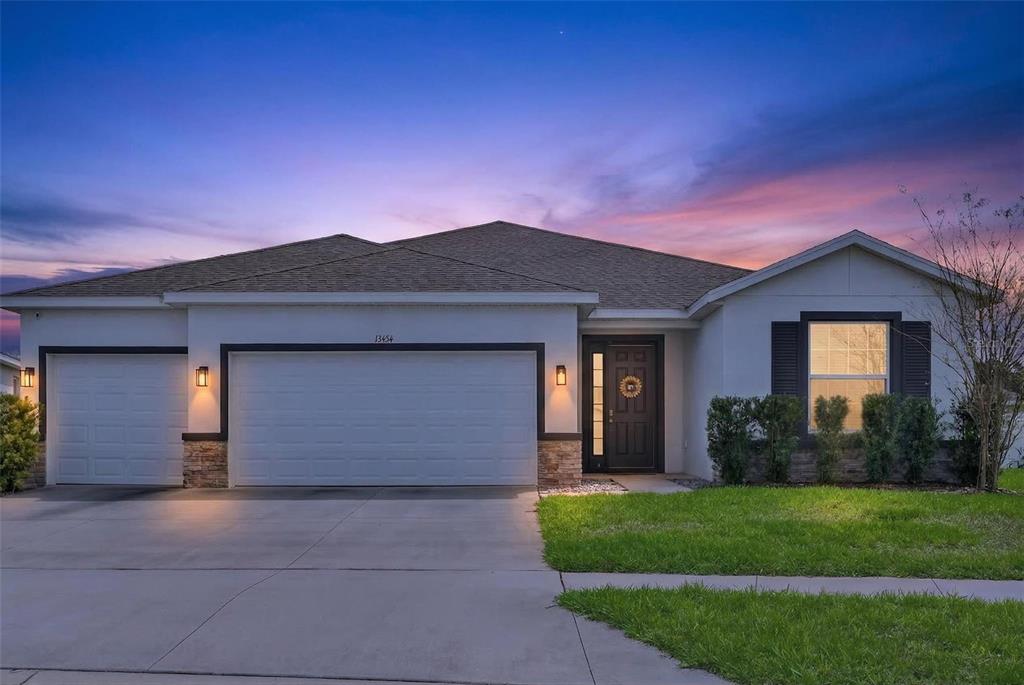 A single-story house with a three-car garage and a dark front door at dusk — 13454 GOLDEN LIME AVENUE, SPRING HILL, FL