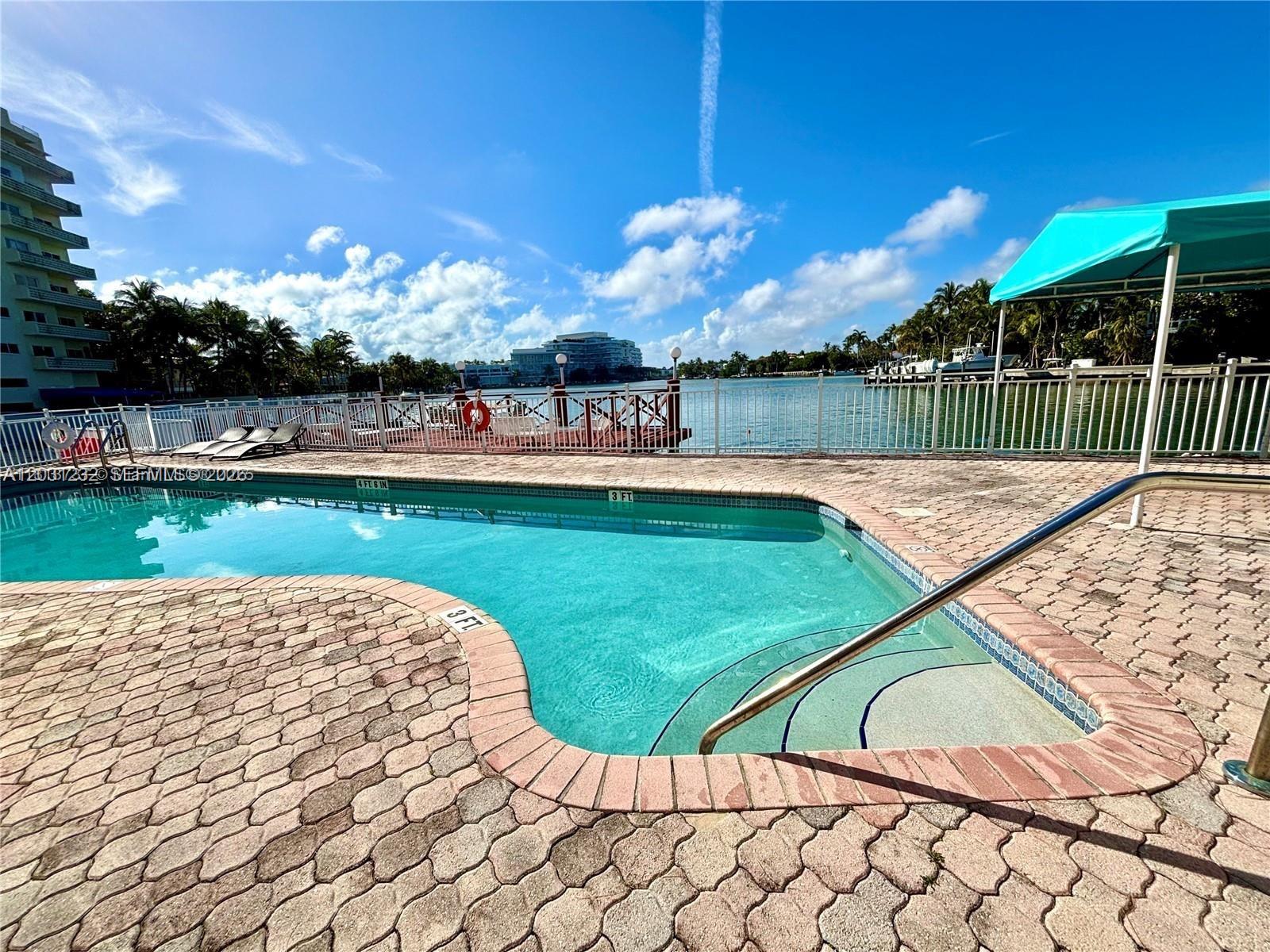 A swimming pool with a brick deck overlooks a bay with a wooden dock — MIAMI BEACH, FL