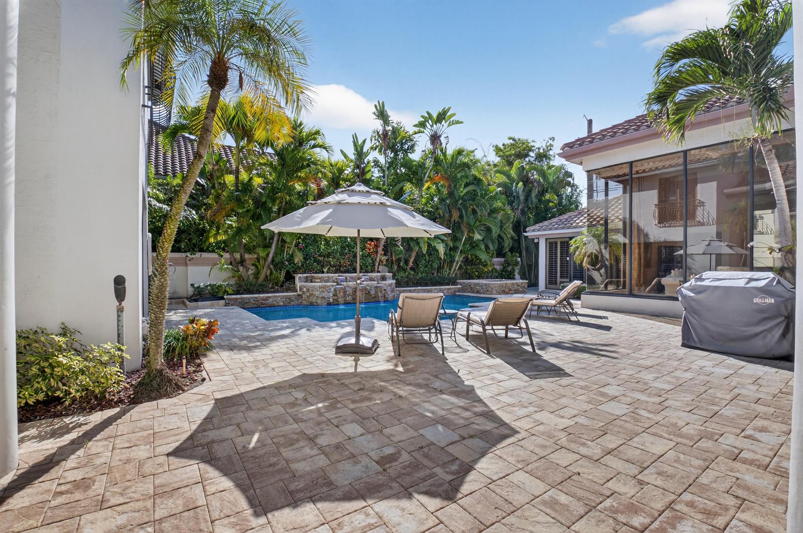 A paved patio features a swimming pool, lounge chairs, an umbrella, and tropical landscaping — 2629 NW 64th Place, Boca Raton FL 33496, BOCA RATON, FL