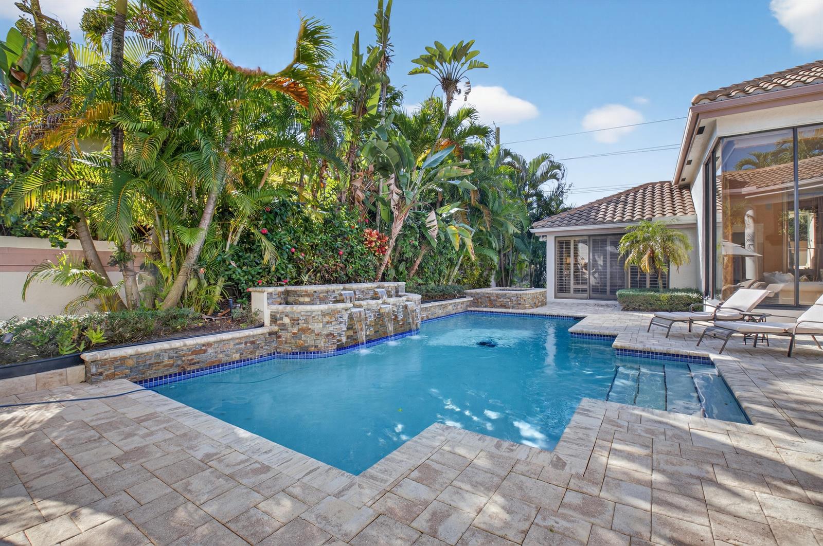 A backyard swimming pool features a stone waterfall, tiled deck, and surrounding tropical landscaping — 2629 NW 64th Place, Boca Raton FL 33496, BOCA RATON, FL
