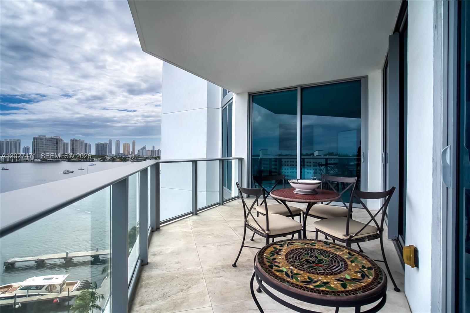 A balcony with tile flooring, patio furniture, and a glass railing overlooking a waterfront cityscape — NORTH MIAMI BEACH, FL