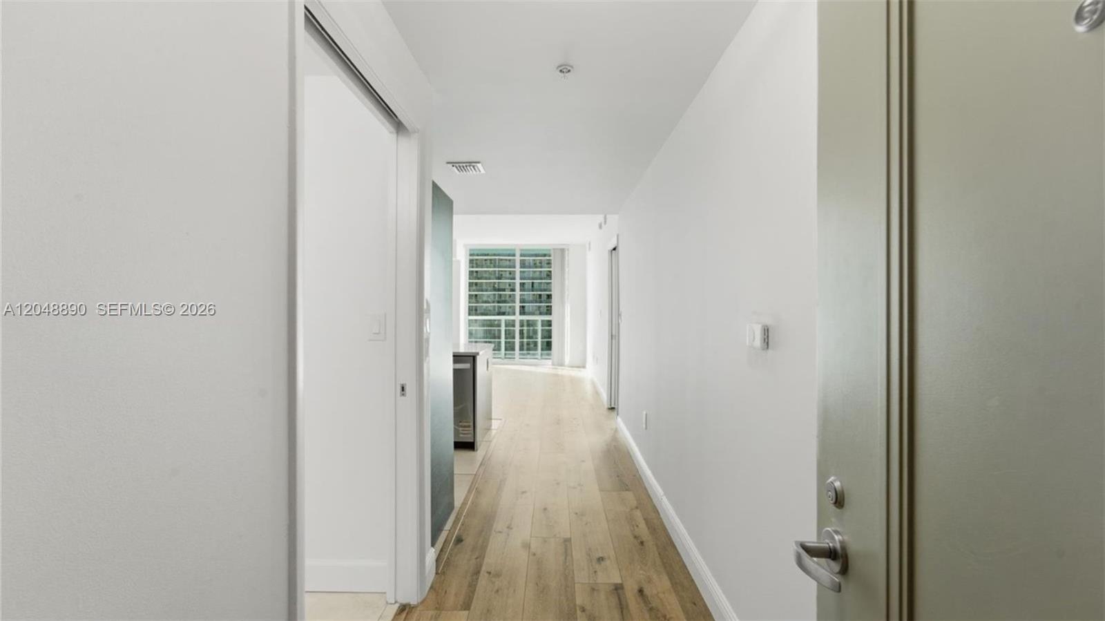 A hallway with light wood flooring leads toward a bright living area with floor-to-ceiling windows — MIAMI, FL