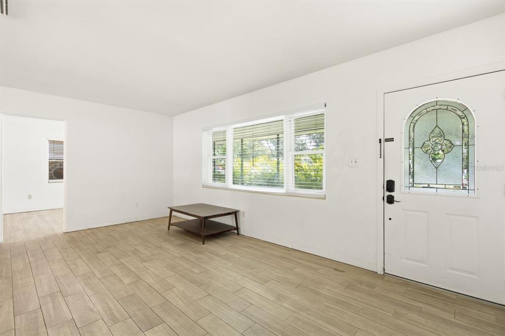 A bright living room features light wood-look tile flooring, white walls, and a decorative front door — 1626 S BETTY LANE, CLEARWATER, FL
