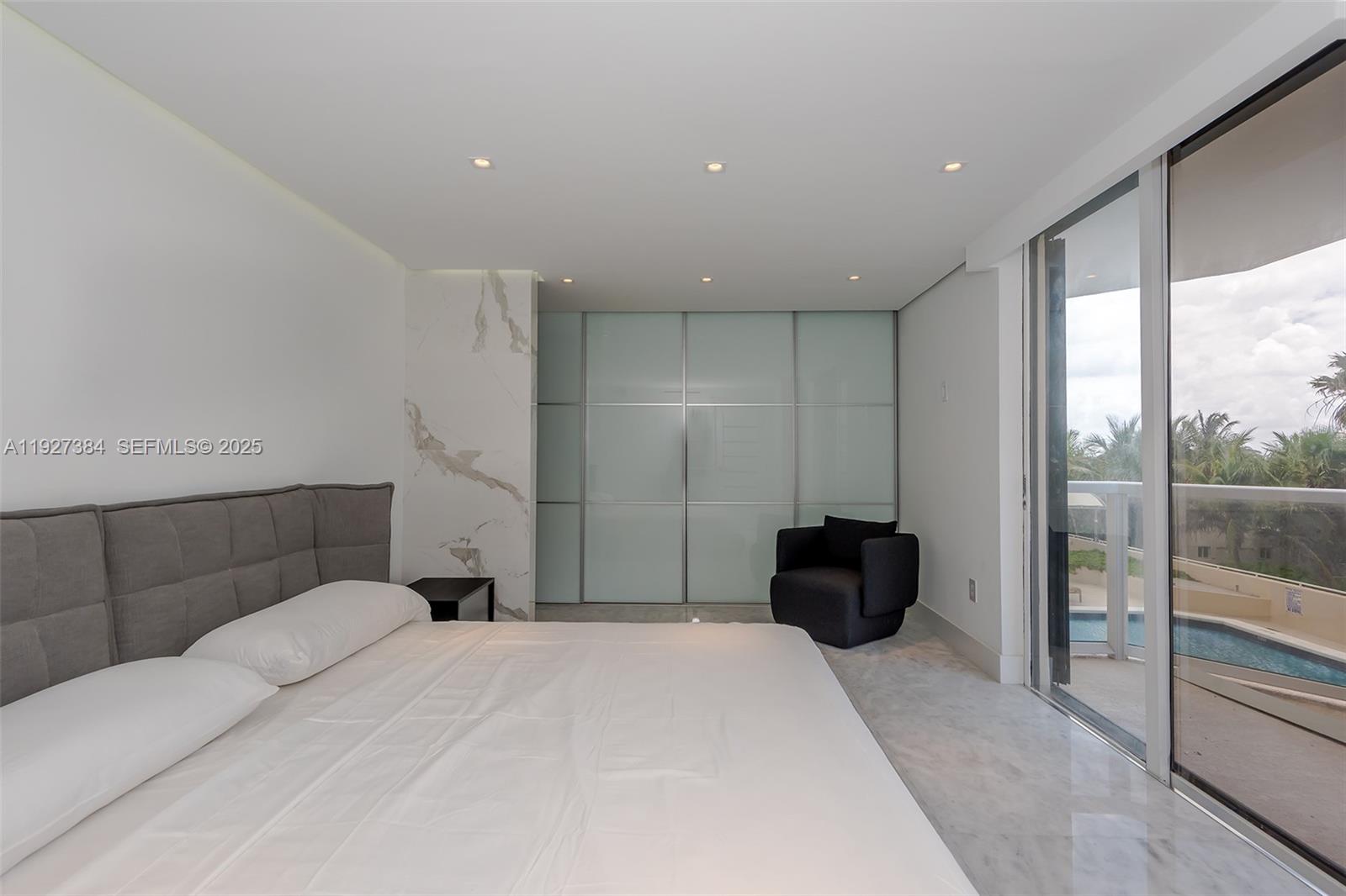 A modern bedroom features a gray tufted headboard, marble accent wall, glass-paneled closet, and balcony access — 6423 Collins Ave # 304, Miami Beach FL 33141, MIAMI BEACH, FL