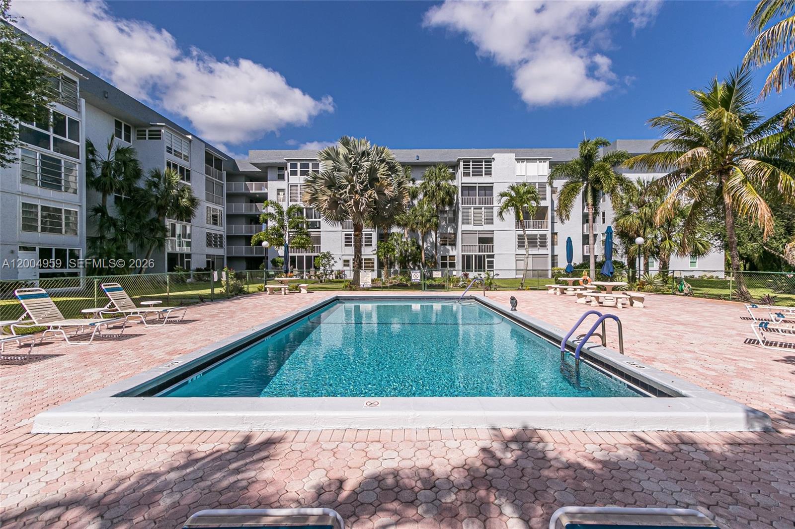 A rectangular swimming pool sits on a paved deck surrounded by palm trees and buildings — 1402 NW 80th Ave # 302, Margate FL 33063, MARGATE, FL