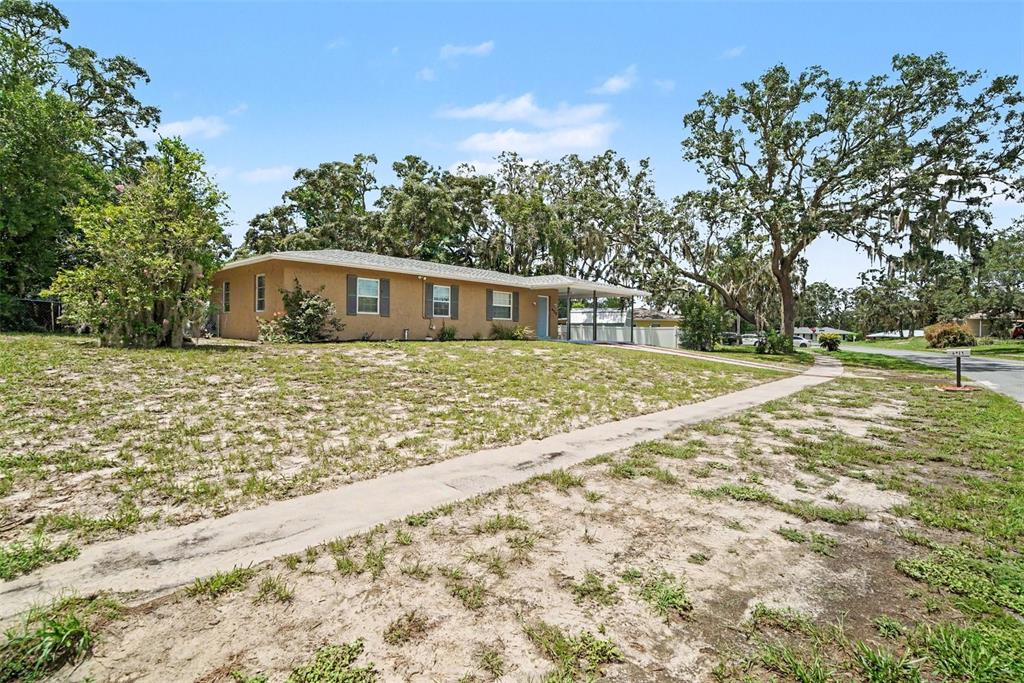 A single-story tan house with a carport sits on a sandy lot with large trees — 6925 TREEHAVEN DRIVE, SPRING HILL, FL