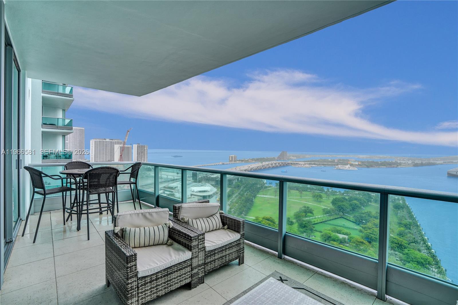 A high-rise balcony features outdoor wicker furniture overlooking a park, a bridge, and the ocean — 900 Biscayne Blvd # 2907, Miami FL 33132, MIAMI, FL