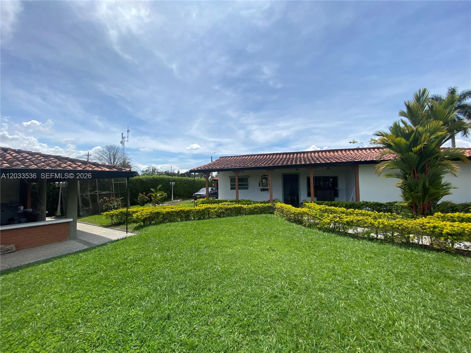 A single-story house with a terracotta tile roof sits behind a manicured lawn and hedge — Casa 21 Barrio Morelia, FL, FL