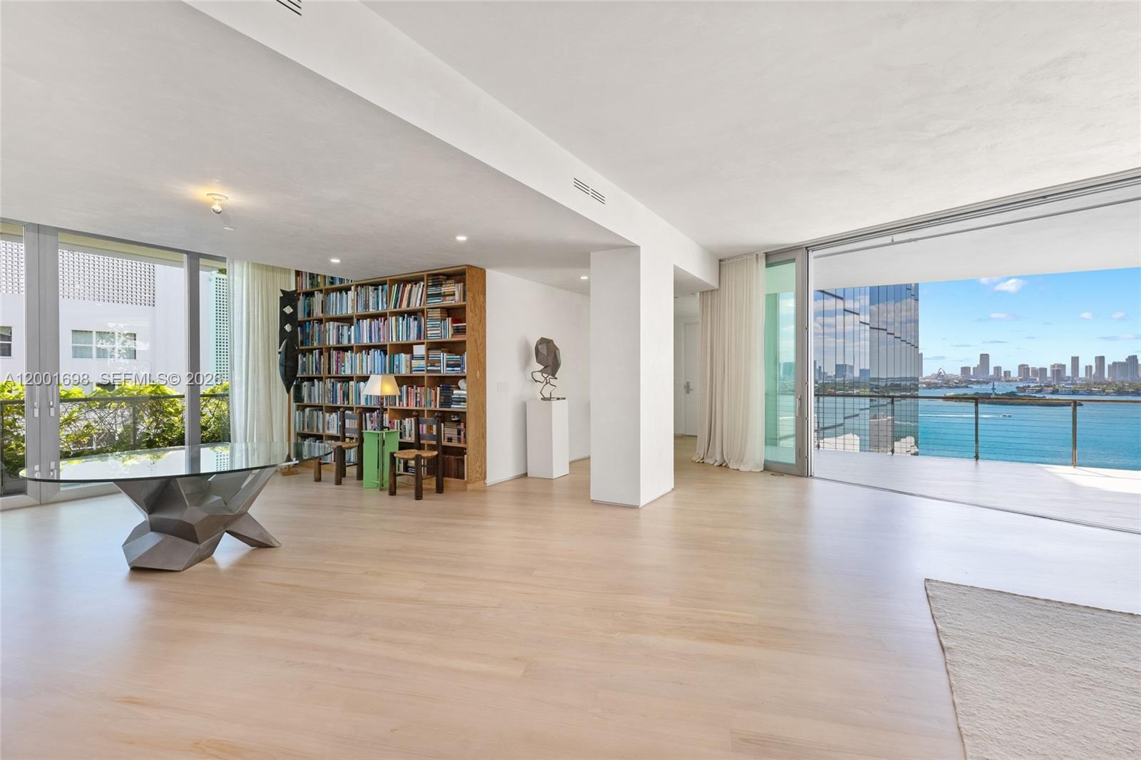 A spacious living area features light wood floors, a large bookshelf, and a balcony view — 1300 Monad Ter # 11E, Miami Beach FL 33139, MIAMI BEACH, FL