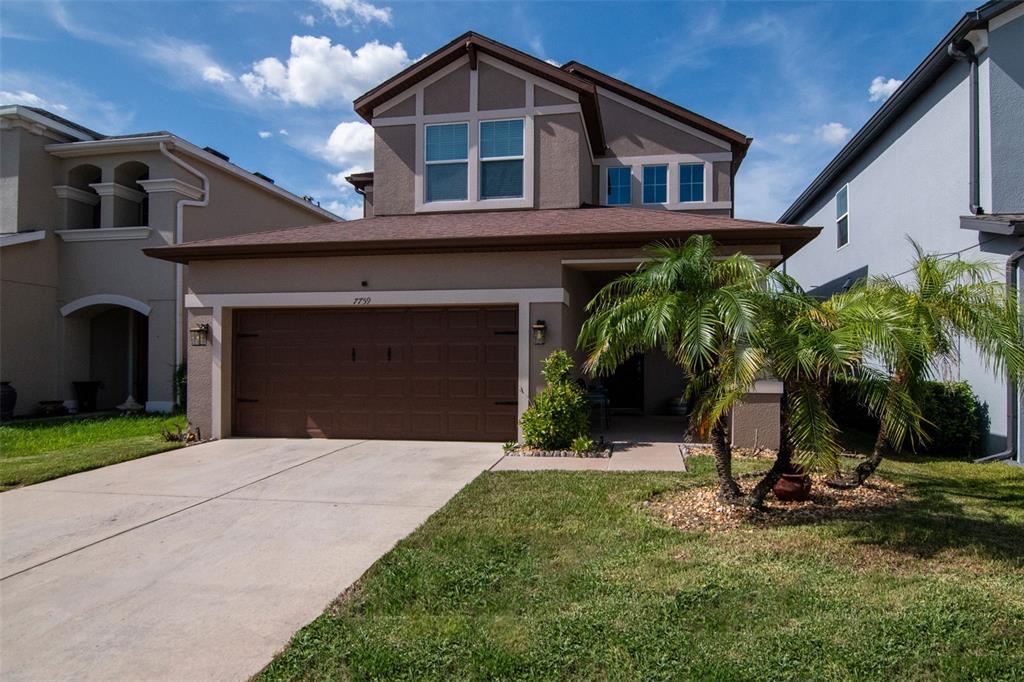 A two-story suburban house with a brown garage door, tan stucco siding, and a palm tree — 7759 WINDCHASE WAY, WESLEY CHAPEL, FL