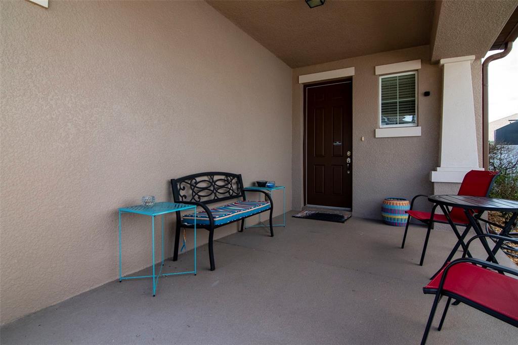 A covered porch features a metal bench, two small tables, and red patio chairs — 7759 WINDCHASE WAY, WESLEY CHAPEL, FL
