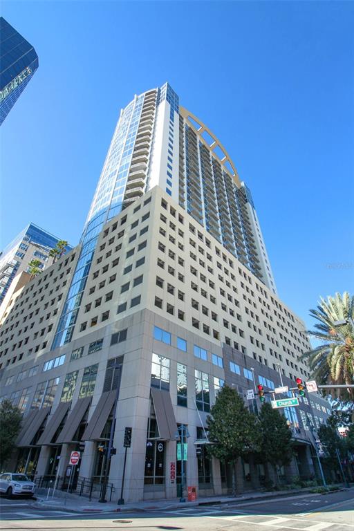 A tall, modern high-rise building with a parking garage base stands on a city street — 155 S COURT AVENUE UNIT 1102, ORLANDO, FL